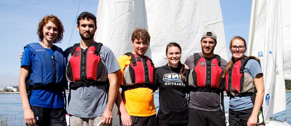 a group of students wearing lifejackets pose in front of sailboats in the water