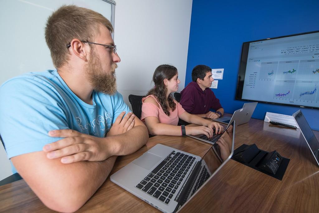 Three students sit with their laptops and review line graphs on a large screen