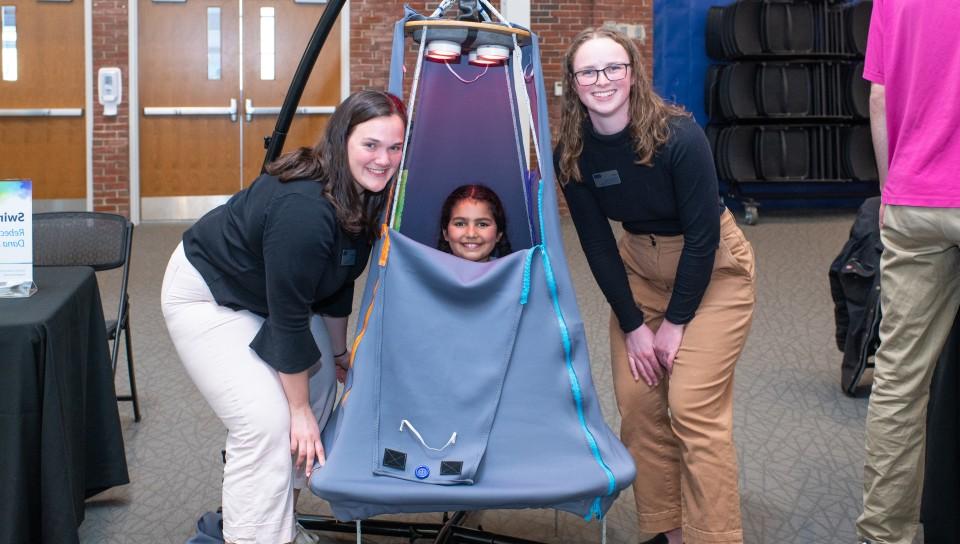 A child sits inside a suspended tent and two students pose around her