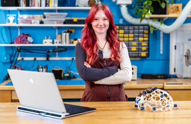 A student stands with arms crossed and smiling at the camera next to a brain wave helmet and laptop