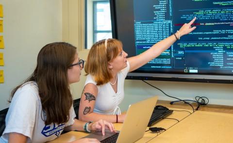 Two students study at a computer with a large TV screen displaying data